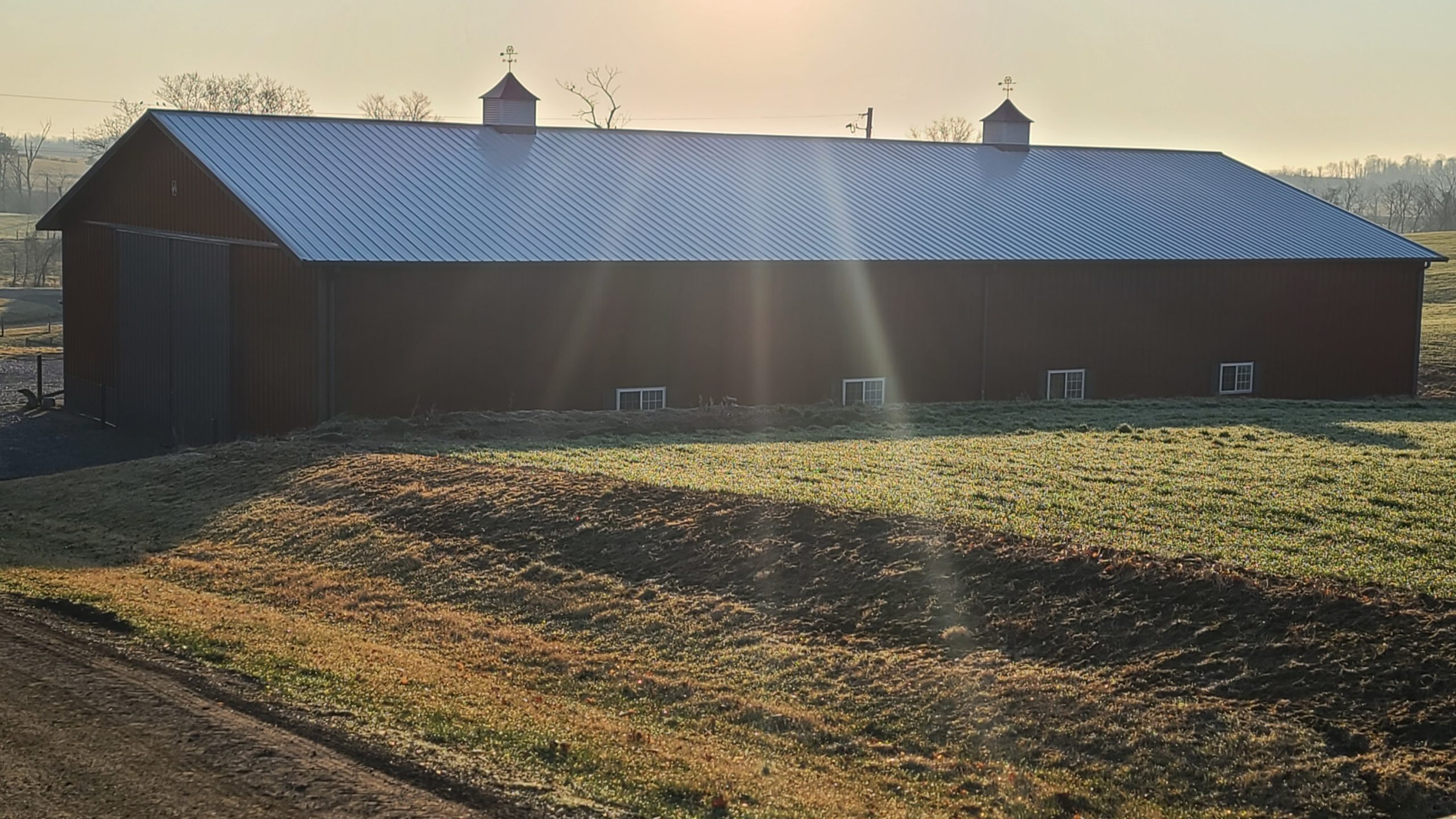 A large barn with a metal roof sits in a sunlit, grassy field, showcasing the quality of its construction. The sun casts long beams across the scene, and two cupolas are visible on the roof. The landscape is serene and slightly foggy in the background.