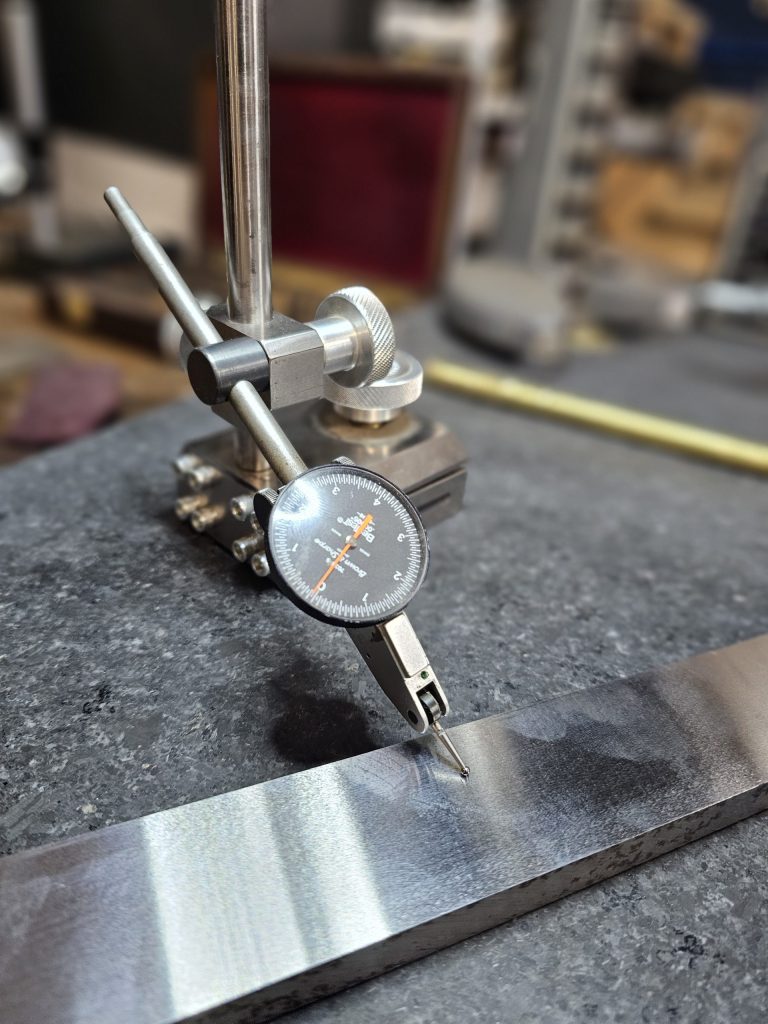 A close-up of a dial indicator mounted on a stand, precisely measuring the surface of a metallic object. The dial shows accurate readings, crucial for machinists assessing material flatness or thickness. Set in Central PA, the blurred background contains workshop tools from Advanced Tooling Solutions.