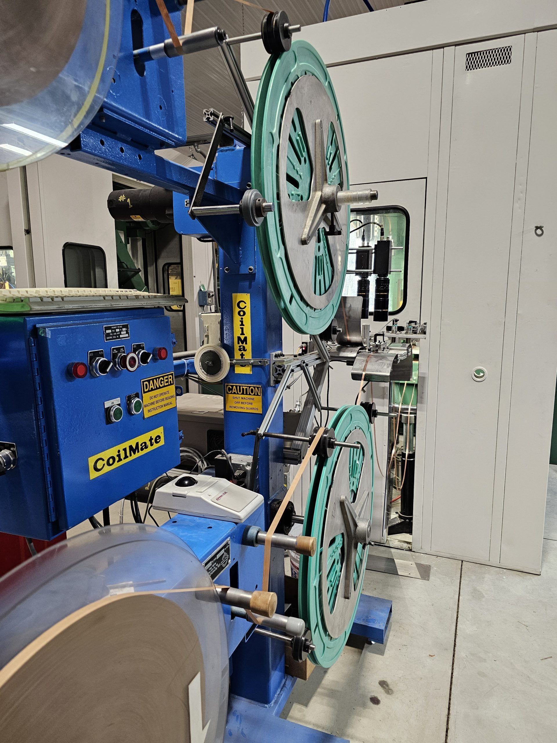 A blue industrial machine with large spinning wheels labeled "CoilMate" stands in a factory environment. It features caution and danger signs, various control panels, and levers, making it essential for machinists. This setup is typical in Central PA's busy manufacturing hubs.