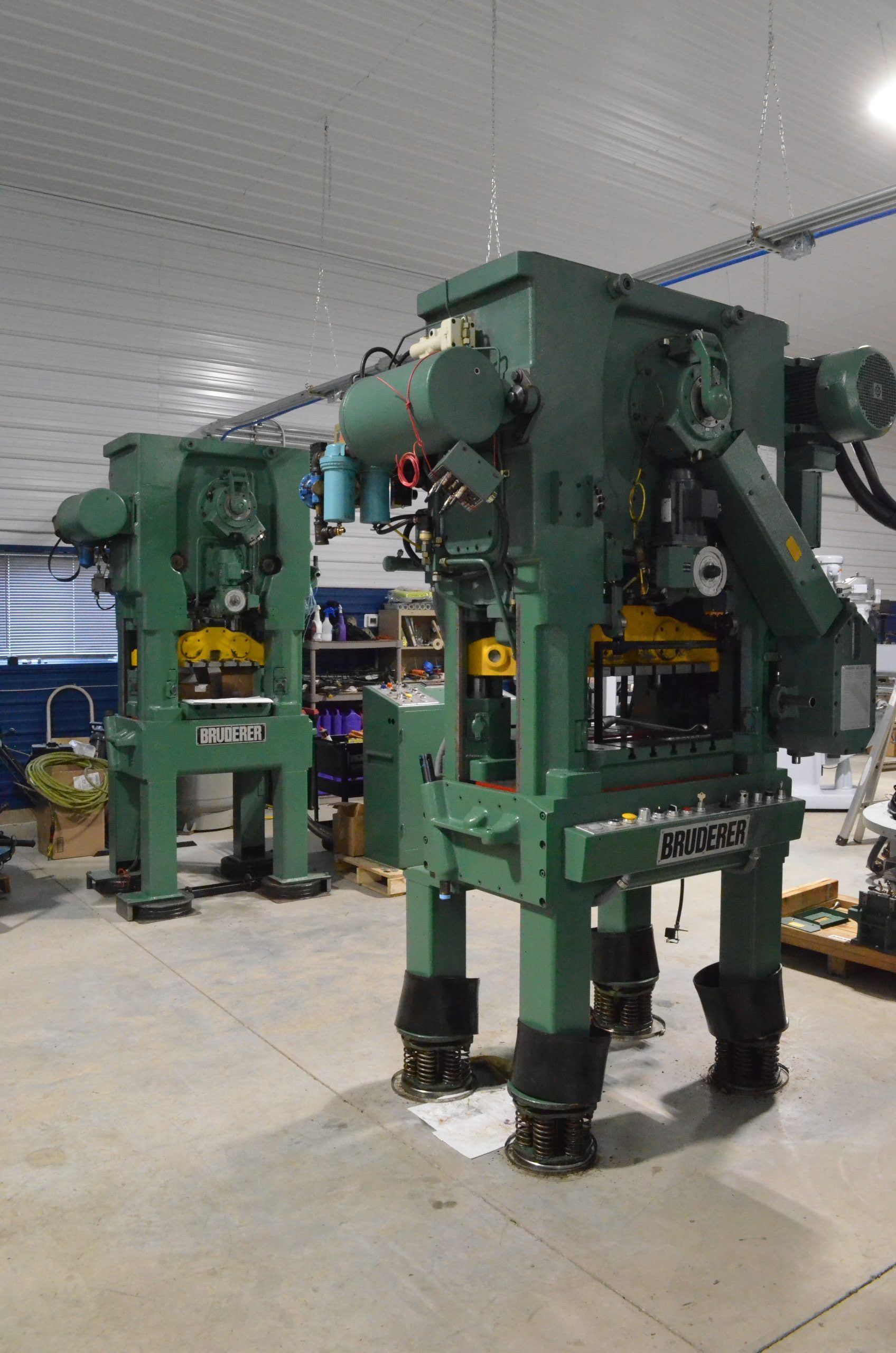 Two green industrial Bruderer punching machines sit side by side in a spacious workshop, ready for machinists to ensure quality production. The machines boast various attached components under high ceilings and bright lights. Tools and equipment are neatly organized throughout the room.