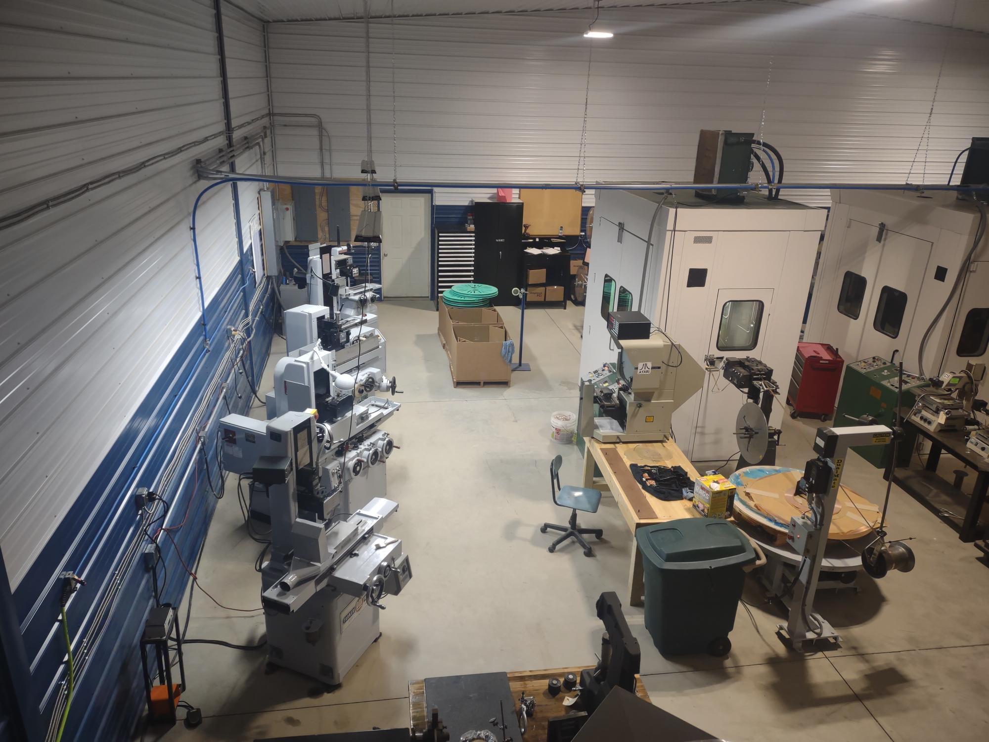 A spacious workshop with industrial machines, such as milling machines and a drill press, is organized along the walls. A central workbench is surrounded by stamping specialists' tools, chairs, and trash bins. The area boasts a high ceiling and excellent overhead lighting.