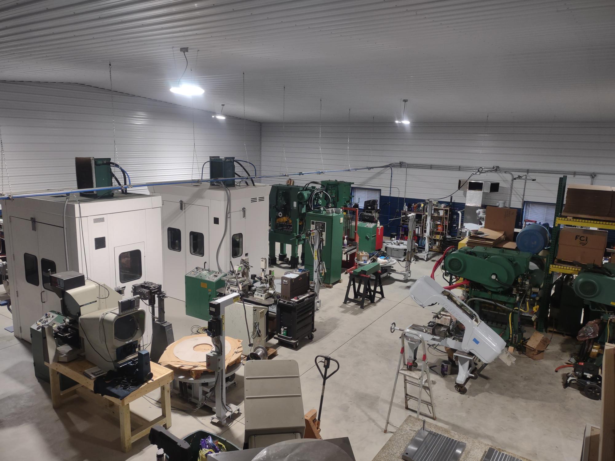 A spacious workshop equipped with green and white industrial machines, worktables, and tools catering to specialists. The room is well-lit with overhead lights, while neatly arranged boxes and supplies sit on shelves.