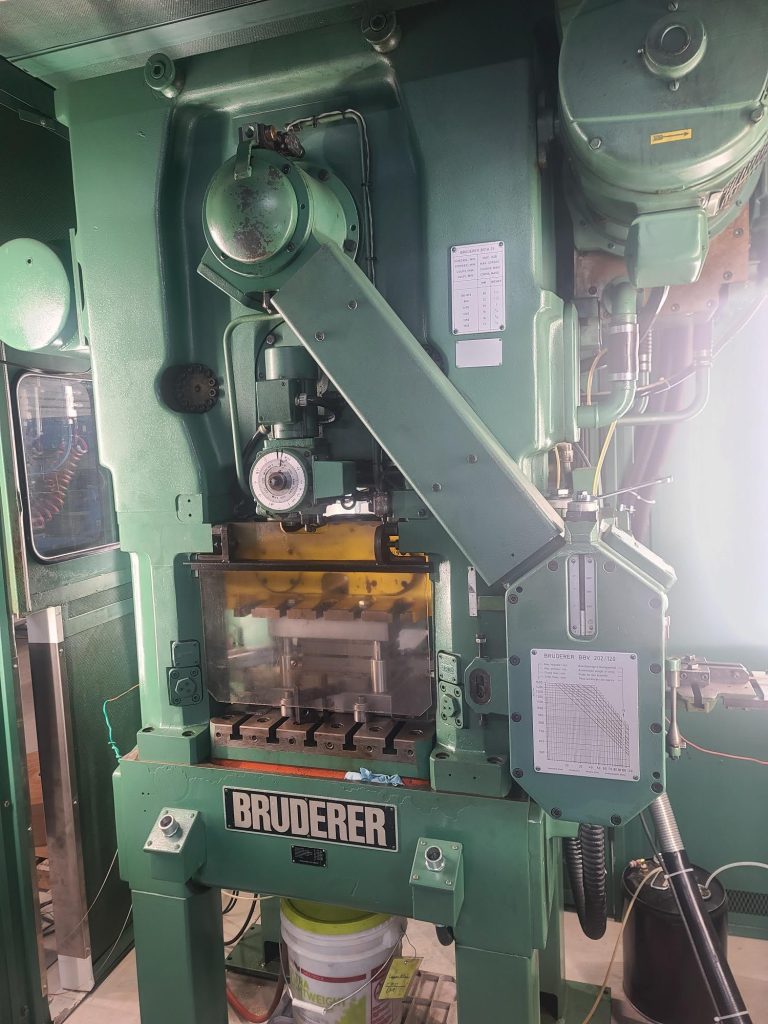 Located in Central PA, a large Bruderer machine press stands in the factory, featuring various levers and controls. Clear sections reveal its internal components, emphasizing quality craftsmanship. Around it, skilled machinists operate additional equipment and tools.