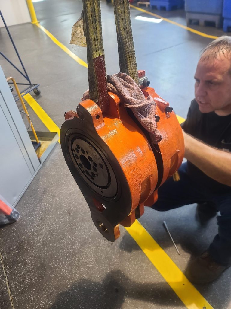 A specialist crouches on a workshop floor, inspecting an orange mechanical part suspended by straps. The area has a gray, polished floor and yellow safety lines. A wrench and a cloth are visible nearby as symbols of advanced tooling solutions ensuring quality in every detail.