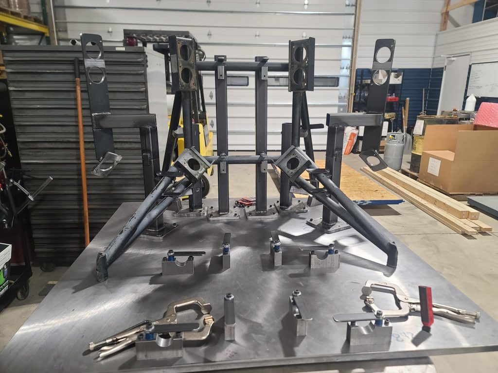 In a workshop in Central PA, a metal assembly jig sits on a table, expertly crafted for holding and aligning components. The background features shelves, cardboard boxes, and an array of equipment that machinists frequently use.