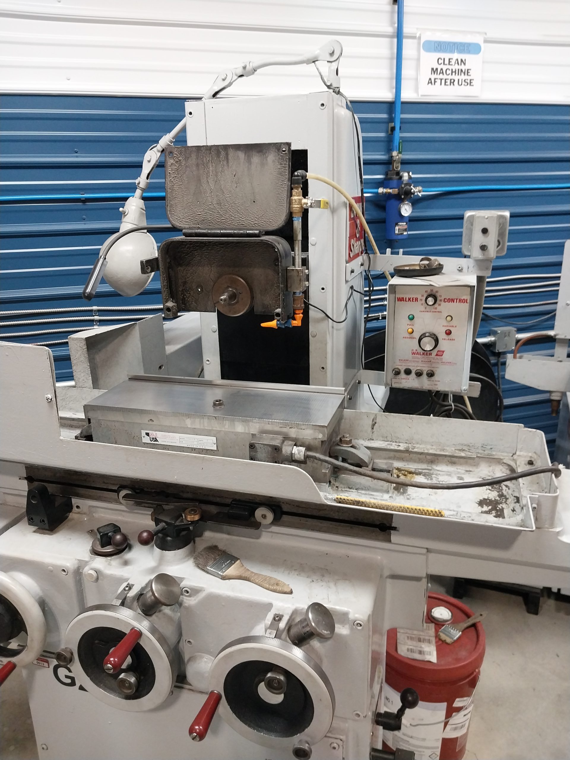 A metal lathe machine equipped with control knobs and levers stands in a workshop, showcasing Advanced Tooling Solutions. There's a tool attached, and a sign above instructs, "Clean machine after use." A blue and white wall serves as the background.