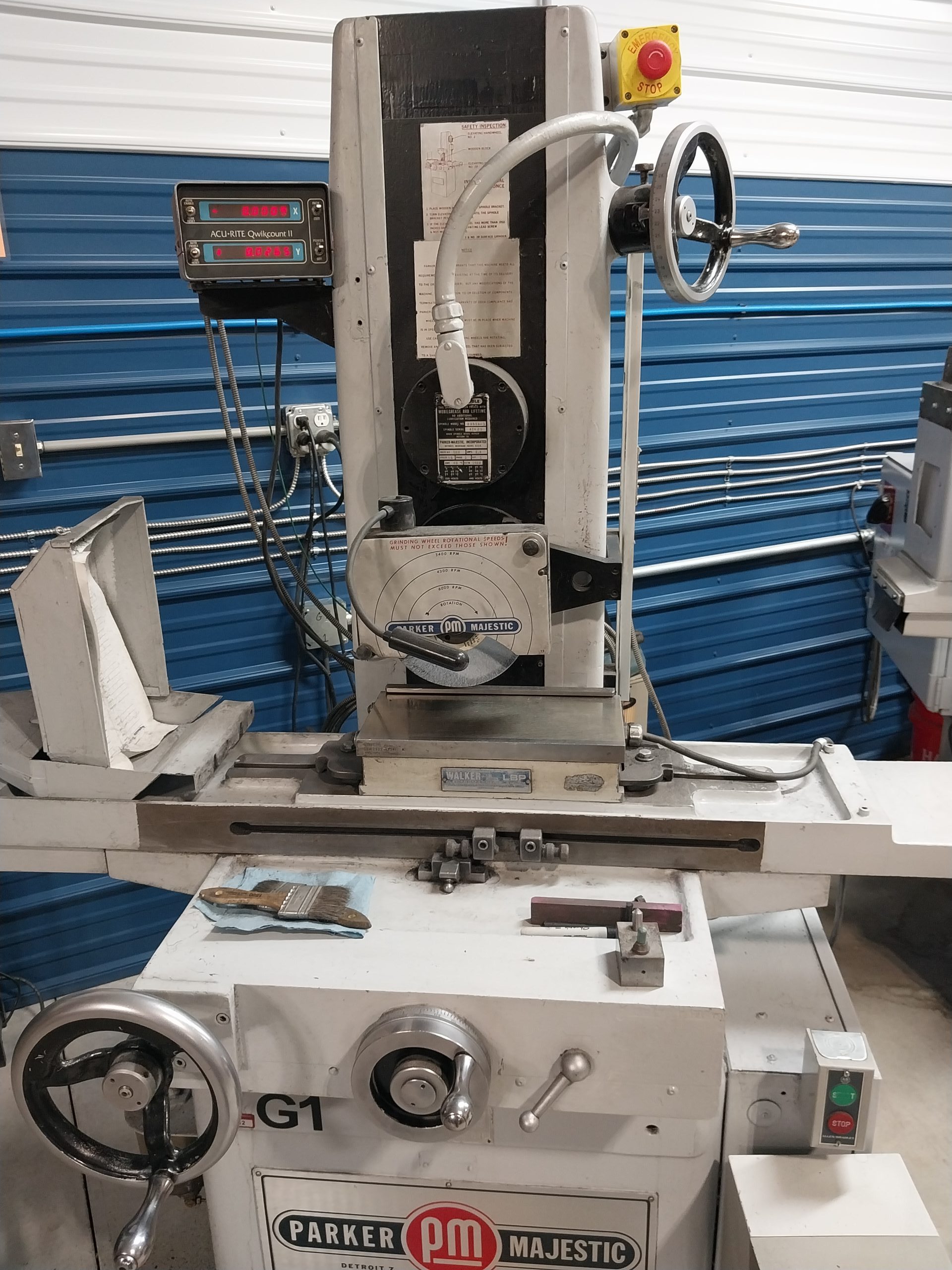 A Parker Majestic grinding machine with a manual control panel and several knobs stands in a workshop with a blue corrugated wall. Known for its quality, it is surrounded by tools and machines on the workbench, representing Advanced Tooling Solutions in Central PA.