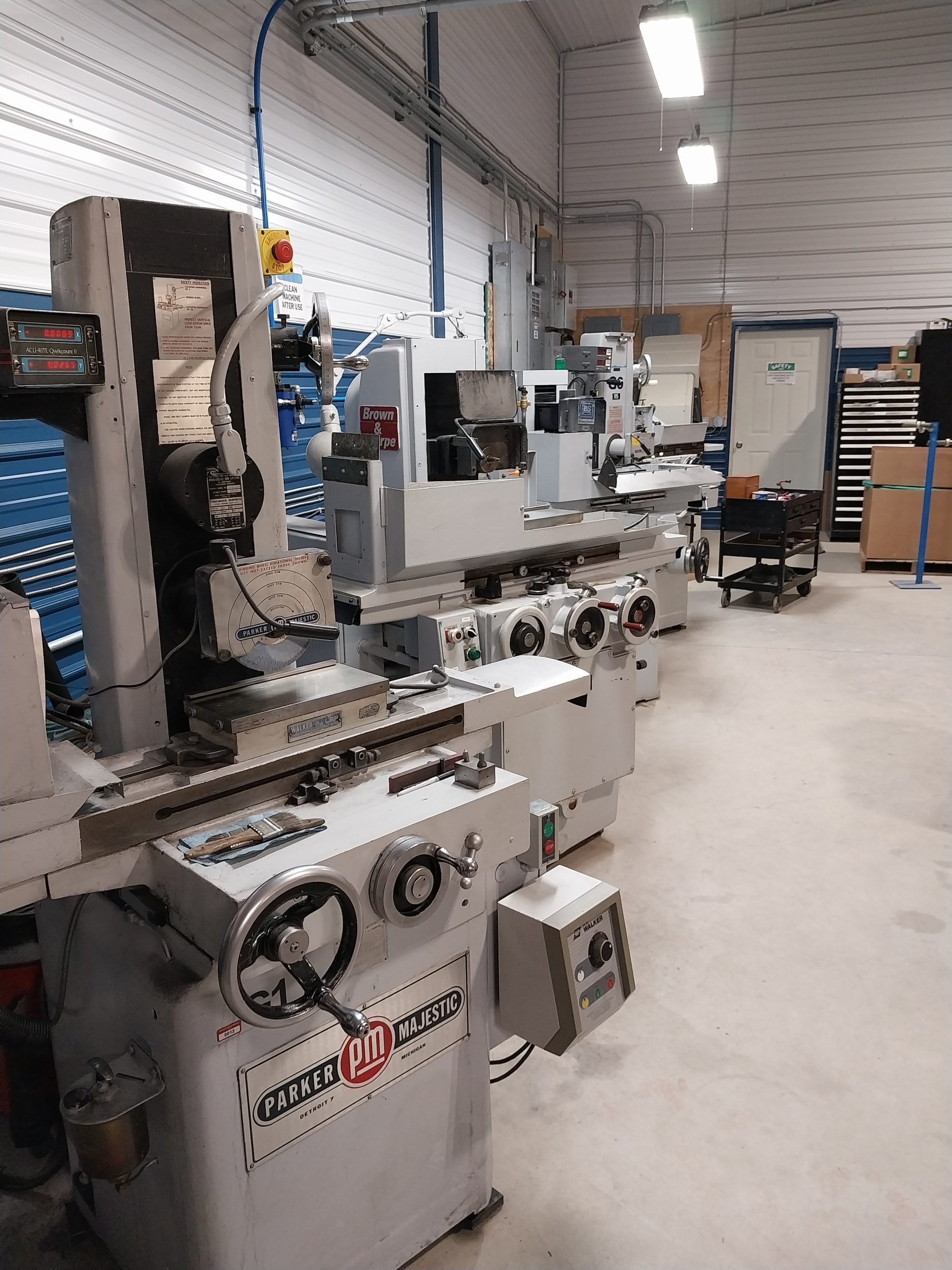 A workshop with several industrial machines, including a surface grinder in the foreground, showcases the quality of tools and equipment. The room has white walls, a high ceiling, and bright lighting. Specialists ensure everything is organized efficiently in the background.