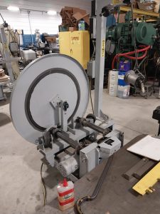 A large industrial machine with a circular component and two horizontal rollers is showcased in a workshop setting, indicating the work of specialists. Various tools and equipment surround the machine. In the foreground, there's a bottle of lubricant and some cables, highlighting quality craftsmanship.