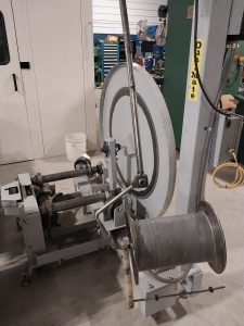 In a workshop setting, a large industrial machine with a metal spool and winding mechanism stands amidst tools and equipment. The vertical frame integrates seamlessly into the environment. Expert machinists utilize Advanced Tooling Solutions to ensure every component meets high quality standards.