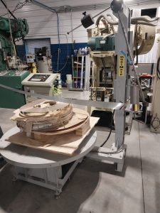 The industrial workshop features advanced tooling solutions with machinery and equipment, including a metalwork machine labeled "RHYMIC." Wooden materials and tools are scattered on a large circular table. The well-lit room showcases various components essential for machinists.