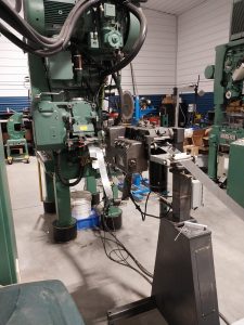 An industrial machine stands in a factory setting, showcasing Advanced Tooling Solutions with precision levers, wires, and components. It's green and black, surrounded by quality equipment on a concrete floor with white and blue walls.