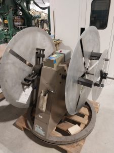 This large industrial machine, featuring two big circular metal plates on a stand, is crucial for stamping operations. Positioned on a wooden pallet, the unit displays switches and controls. The scene’s backdrop includes a white wall and various equipment, reminiscent of Central PA machinists' workspaces.