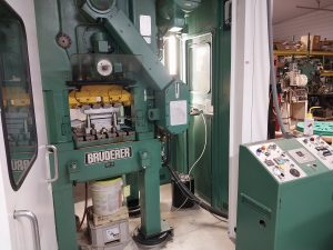 A large green Bruderer industrial stamping machine stands in a workshop, where skilled machinists navigate the control panel with precision. Surrounded by various tools and equipment, this machine operates at the core of their trade.