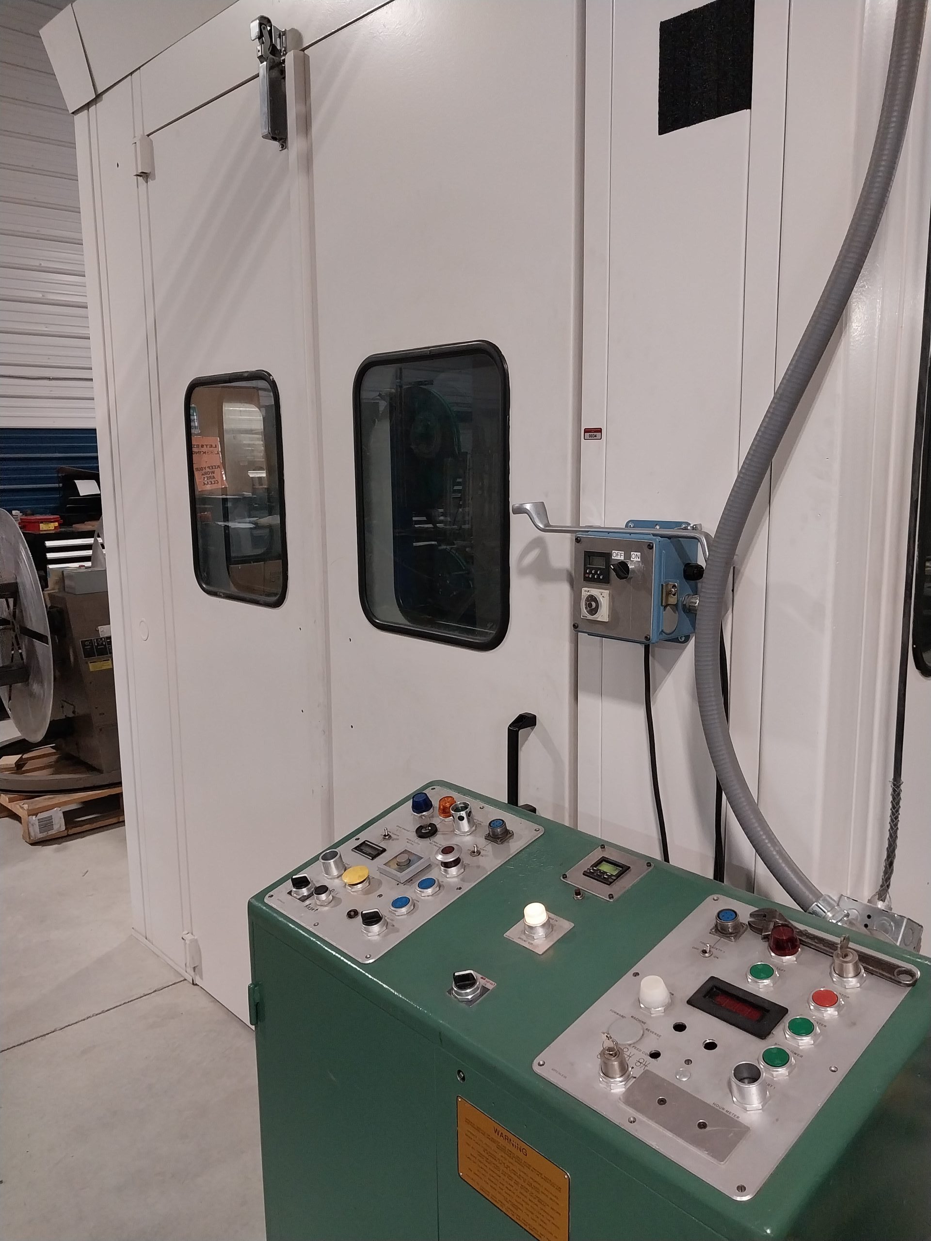 A green control panel with various buttons and switches stands before a large industrial white enclosure with two windows, likely used by specialists. The setup is situated in a spacious facility, with some machinery visible in the background.