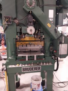 A large green industrial machine, labeled "BRUDERER," stands in a workshop bustling with machinists. It features a control panel, wires, and a central stamping mechanism. A bucket labeled "Bruderer" sits on the ground in front of it, embodying the precision and skill found in Central PA's workshops.