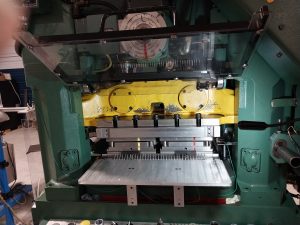 A close-up of stamping machinery with a green and yellow exterior. The machine features metal components and a gauge at the top, set in a factory environment in Central PA, with tools and equipment visible in the background.