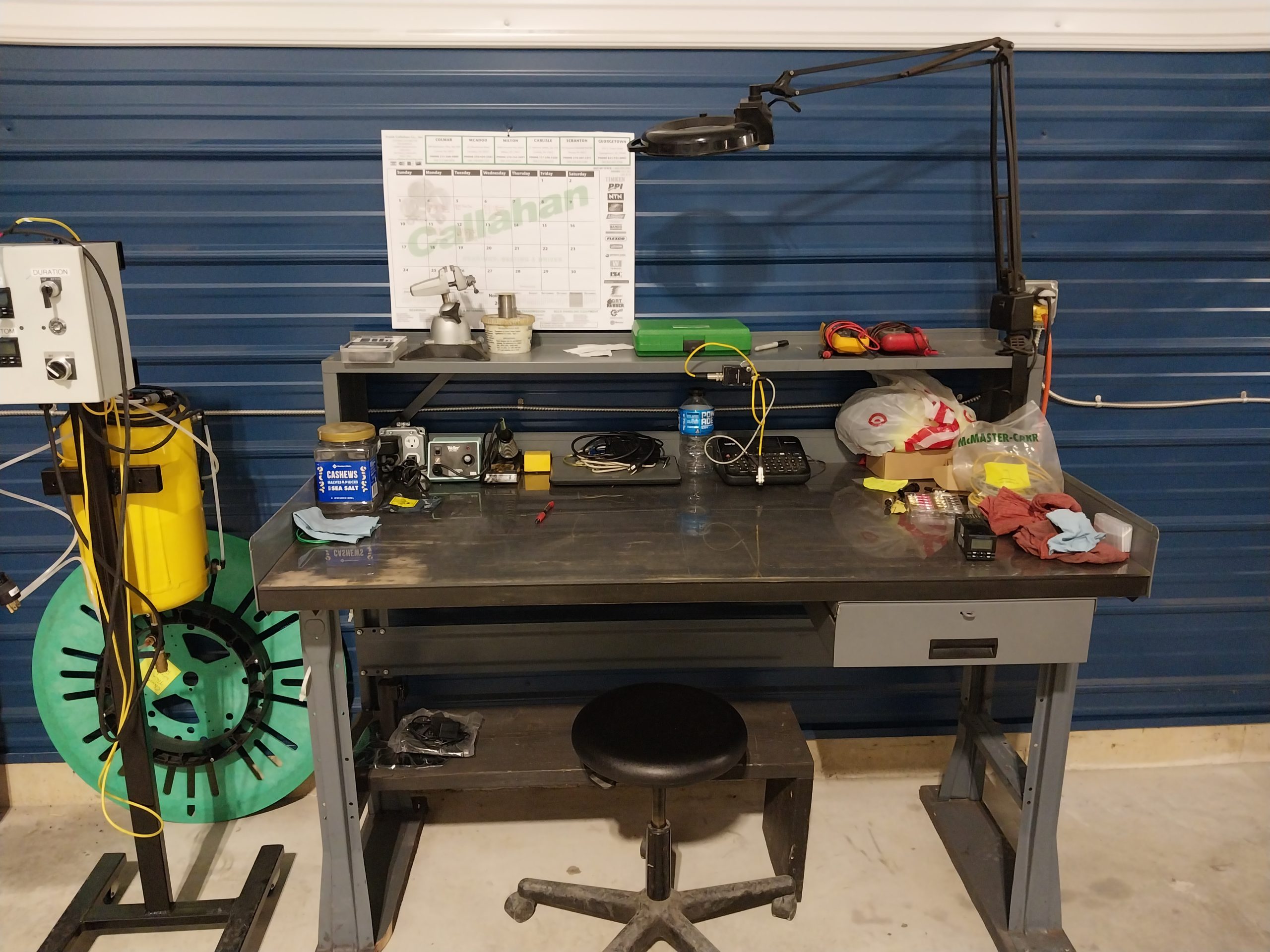 A machinist's cluttered workbench against a blue wall, laden with tools, electronics, a calendar, and a desk lamp. On the left sits a yellow air compressor and green hose reel, while a black stool stands ready in front of the bench.