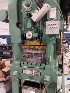 A large green industrial machine with various gears, levers, and a control panel stands ready for skilled machinists and specialists. It features a transparent safety cover, proudly displaying the brand name "BRUDERER," amid boxes and tools in Central PA.