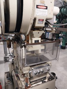 A large industrial stamping machine with a warning sign, featuring various controls and mechanisms. It has a transparent safety shield at its front, situated in a workshop with machinists operating nearby and another machine visible in the background.