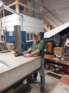 A man in a green t-shirt and cap is smiling as he skillfully operates a metal tool at his workbench in a cluttered workshop. Surrounded by shelves, boxes, and machinery, his expertise as a machinist ensures quality workmanship.