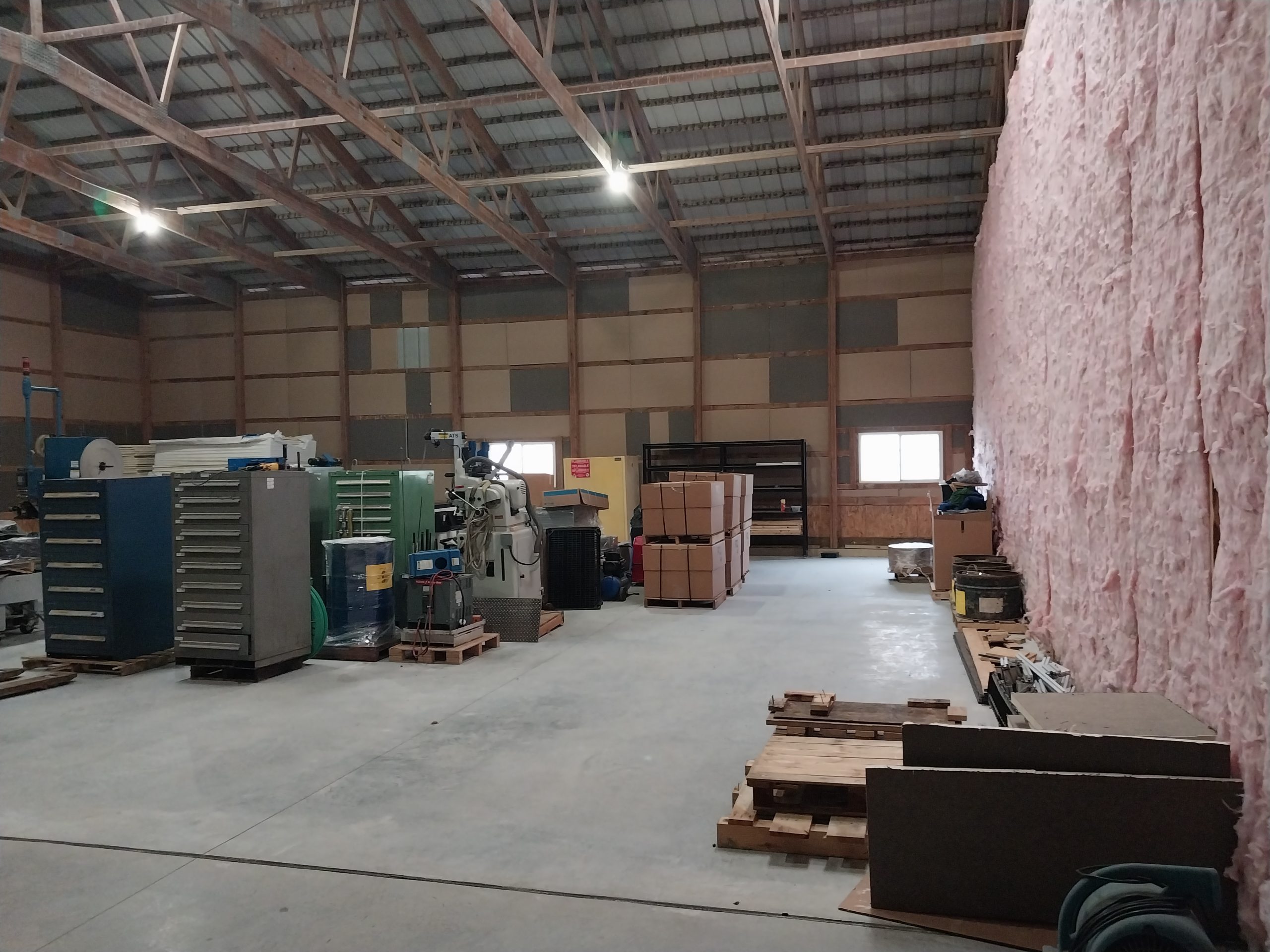 The spacious industrial warehouse features high ceilings and exposed beams, partially insulated with pink material. One side houses pallets and assorted equipment, while large cardboard boxes are stacked in the center. Specialists utilize advanced tooling solutions to optimize operations within this dynamic space.