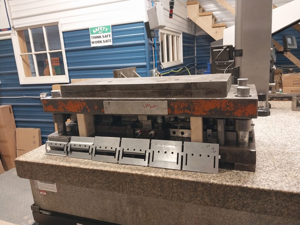 In an industrial workshop, machinists work with a metal stamping die set on a granite table. The die showcases several precise cutouts and has an orange, slightly worn surface. The background features a window, safety signs, and various equipment.