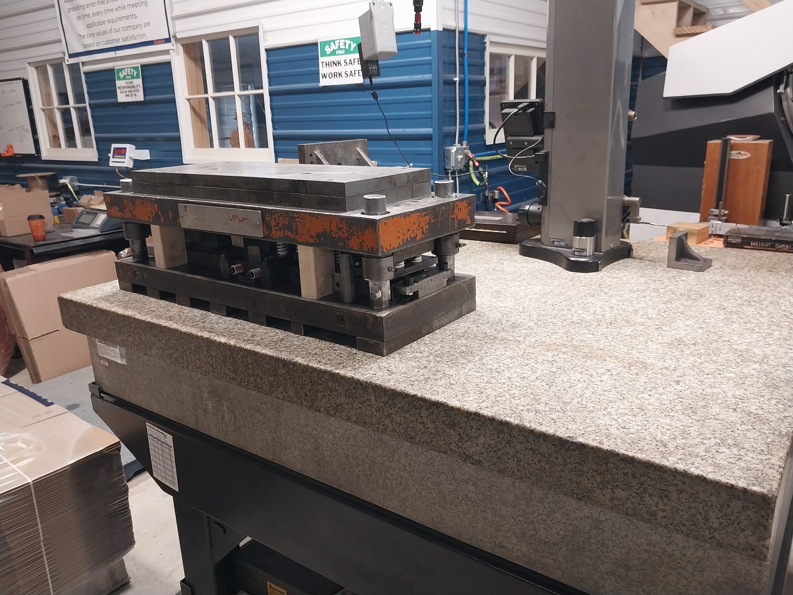 The image depicts industrial machinery on a large granite workbench. This equipment, possibly a metal press, is part of the advanced tooling solutions offered by specialists in Central PA. A blue and white structure stands in the background, with prominent signs emphasizing safety.