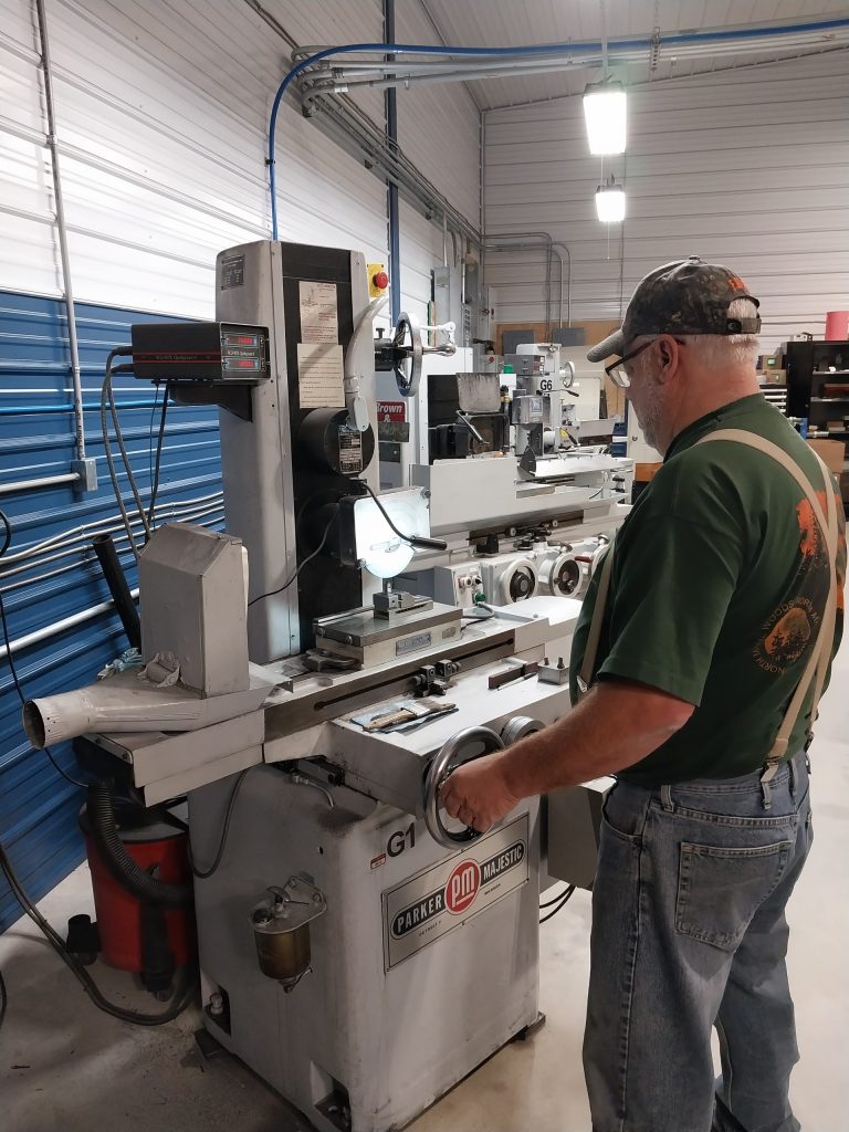 A man in a green shirt and cap operates a Parker PM Machine in a workshop with white and blue striped walls, illuminated by overhead lights. Various tools and equipment are visible, showcasing advanced tooling solutions.