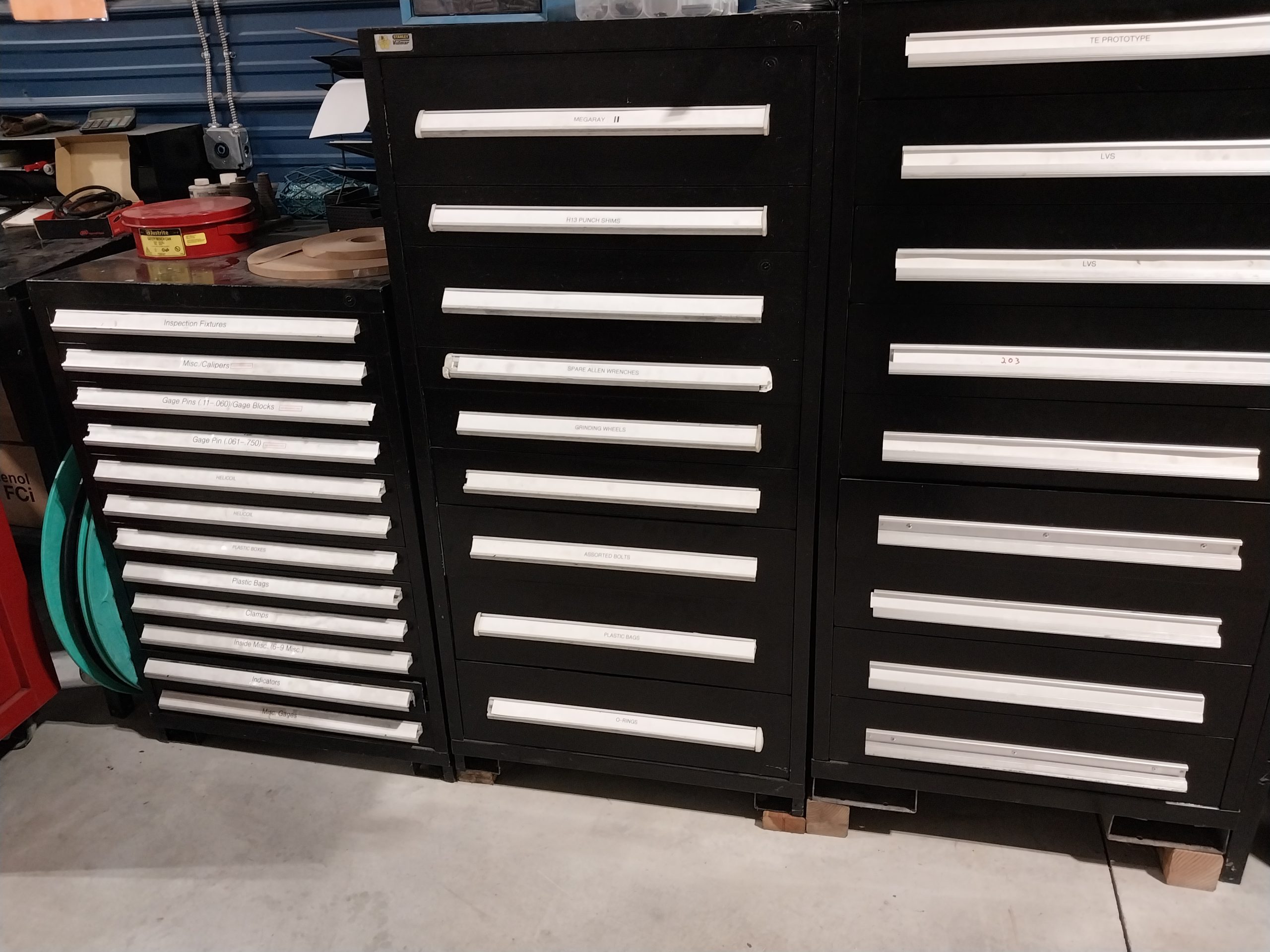 A row of black tool cabinets with white-labeled drawers stands on a light-colored floor in a workshop, showcasing advanced tooling solutions. The cabinets are filled with tools and supplies, while a red toolbox is partially visible on the left, catering to machinists.