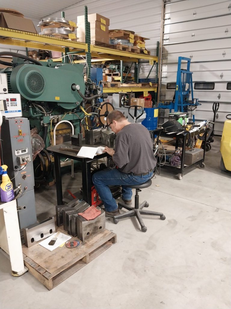 A man sits on a stool in a workshop, meticulously focused on paperwork at a small desk. Around him are various machines and tools, including specialists' equipment like a large industrial stamping machine and shelving units filled with supplies.