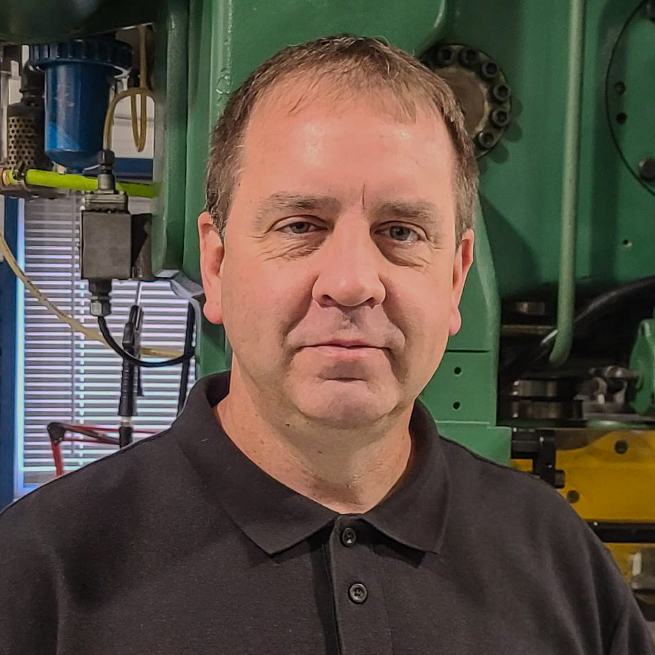 A man in a black polo shirt stands before industrial machinery at Advanced Tooling Solutions, looking at the camera with a neutral expression.