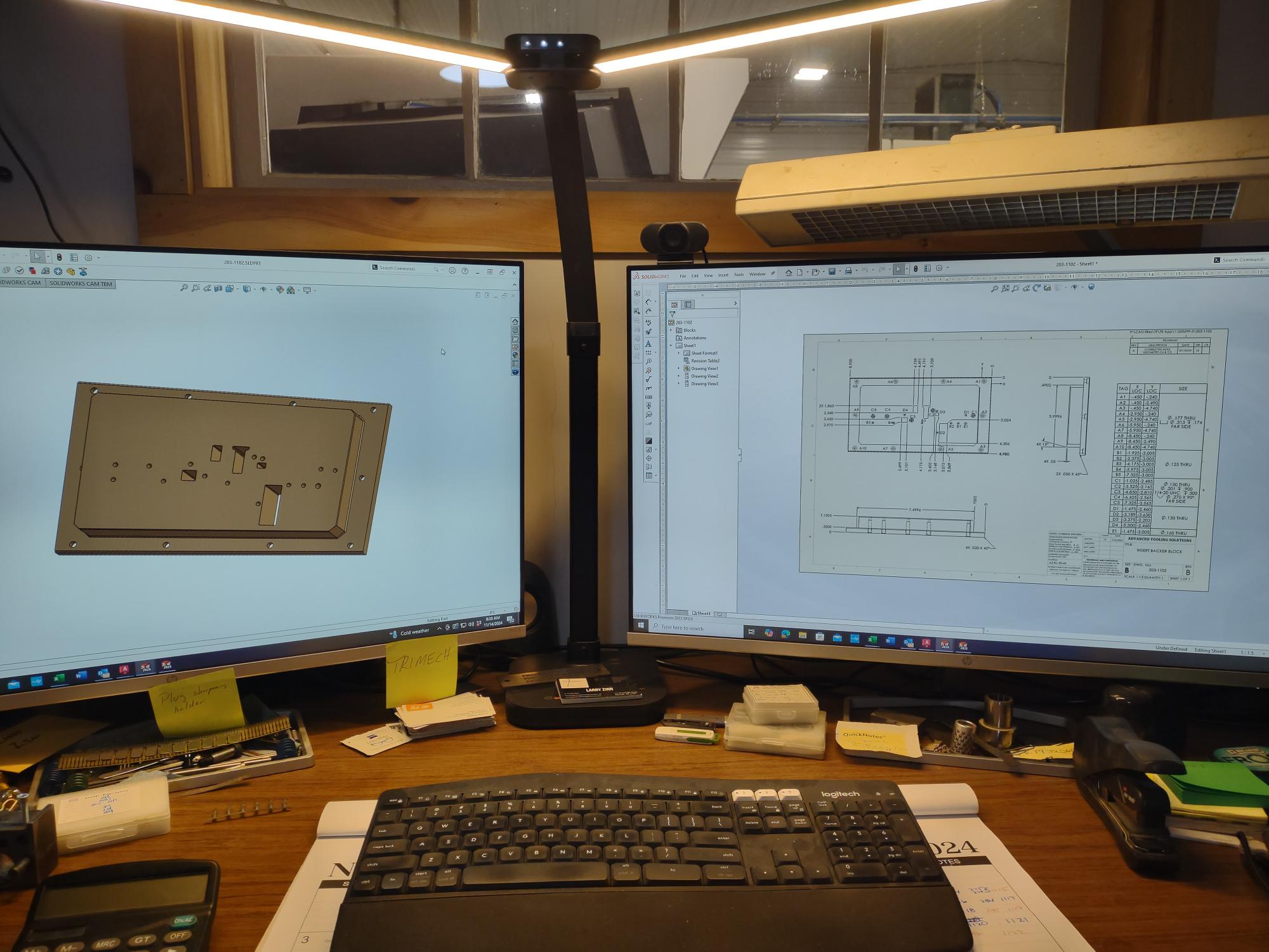 A workspace featuring two monitors displaying a 3D model and a technical drawing. A keyboard sits in front, accompanied by various office supplies like pens and sticky notes. With Advanced Tooling Solutions at hand, a desk lamp perfectly illuminates the area from above, enhancing quality productivity.