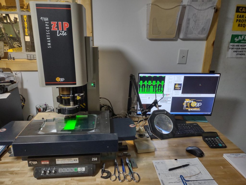 A workspace tailored for specialists showcases a SmartScope ZIP Lite machine beside a computer monitor with imaging software. The desk is equipped with Advanced Tooling Solutions, including a keyboard, magnifying glass, tools, and paperwork. Various measurement instruments are arranged neatly.