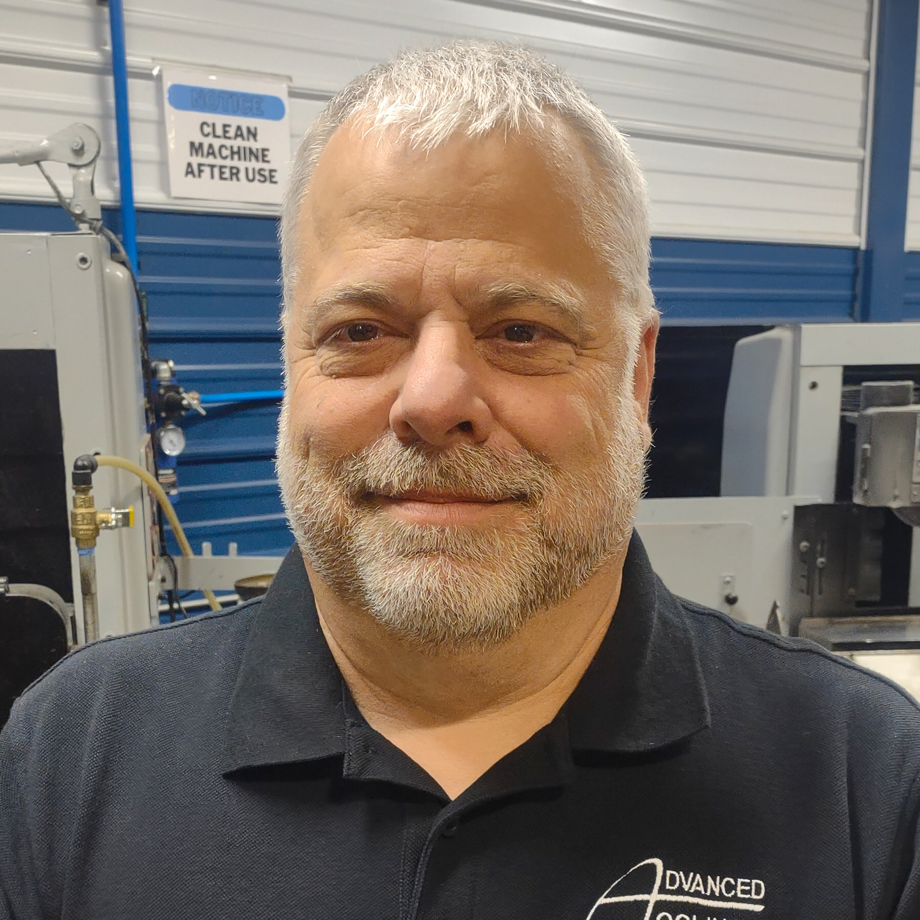 A man with a gray beard smiles while standing in an industrial setting. He is wearing a black polo shirt with the Advanced Tooling Solutions logo. Machinery and a sign that reads "Clean Machine After Use" are visible, highlighting the commitment to quality in this stamping environment.