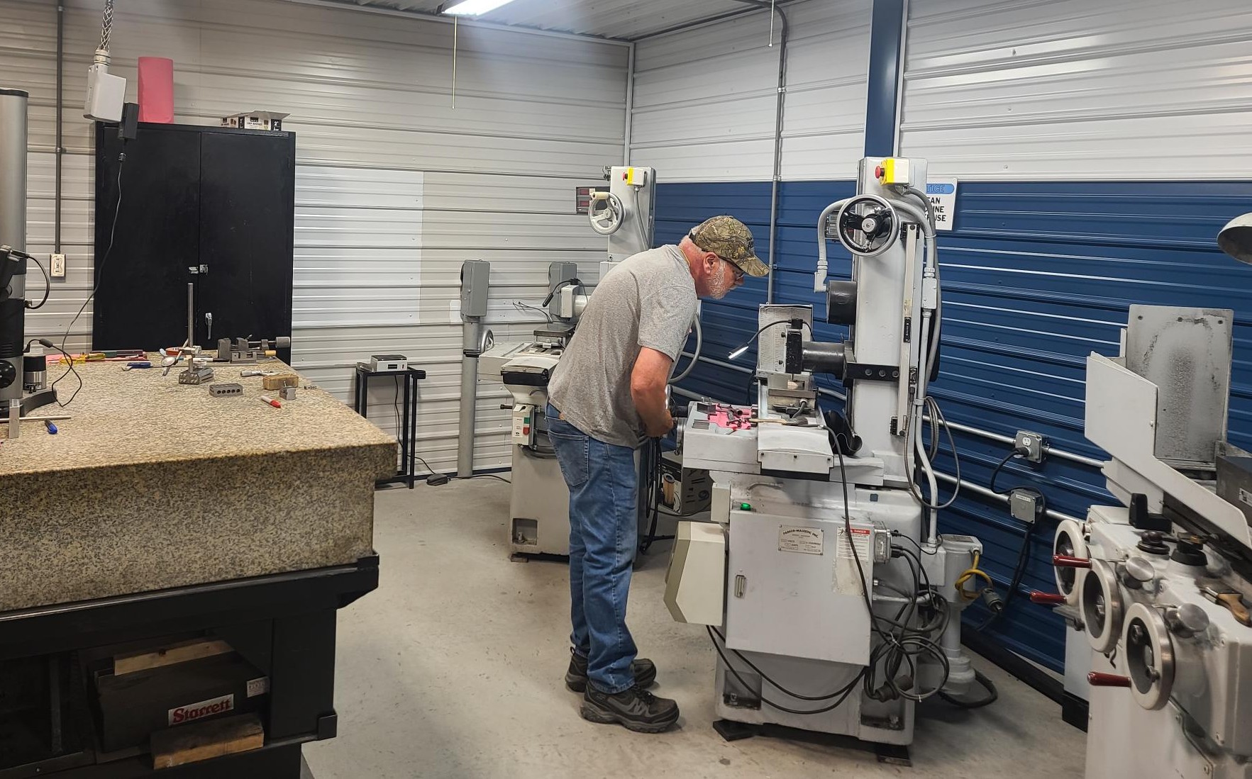 A man in a gray shirt, blue jeans, and a camouflage cap operates machinery in a workshop offering metal stamping services, with white and blue paneled walls, surrounded by equipment, tools, and granite-topped tables.