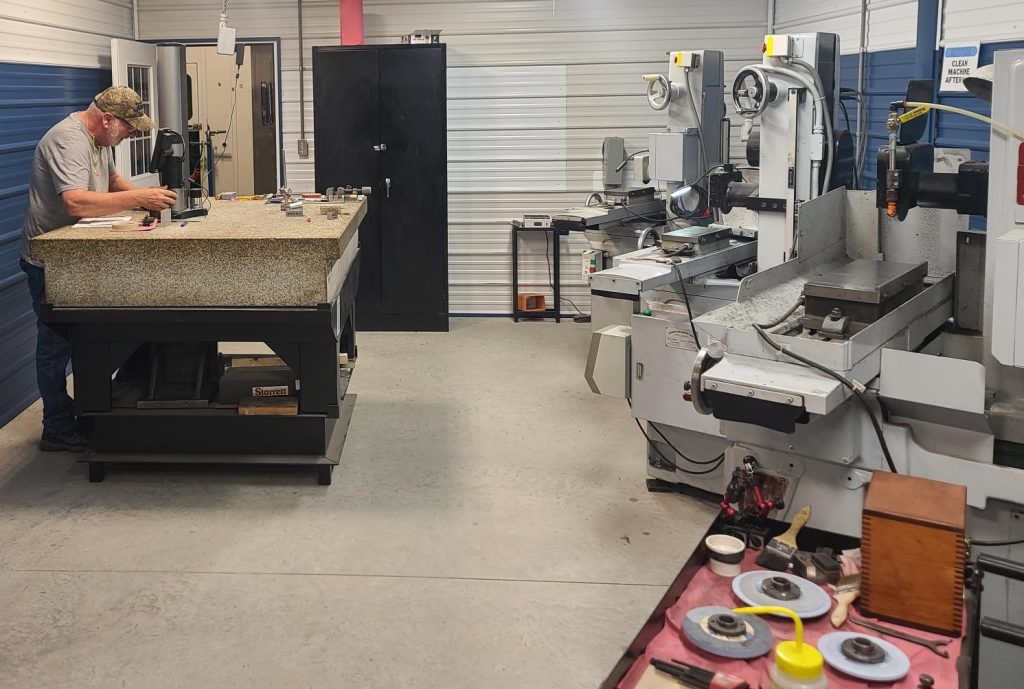 A man wearing a cap works at a granite-topped table in a clean, organized workshop offering metal stamping services, surrounded by precision tools and large CNC machines lining the walls.