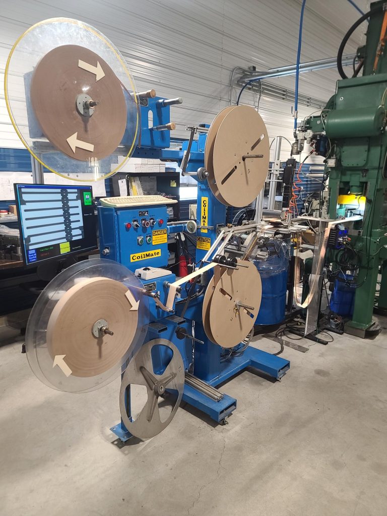 A blue industrial machine with large spools arranged vertically and horizontally, feeding strips of material through rollers—ideal for metal stamping services. Computer monitors and various controls are visible in a workshop setting.